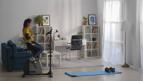 woman-is-doing-sport-in-living-room-of-her-modern-apartment-sitting-on-exercise-bike-and-using-smartphone-healthy-lifestyle-sporty-lady